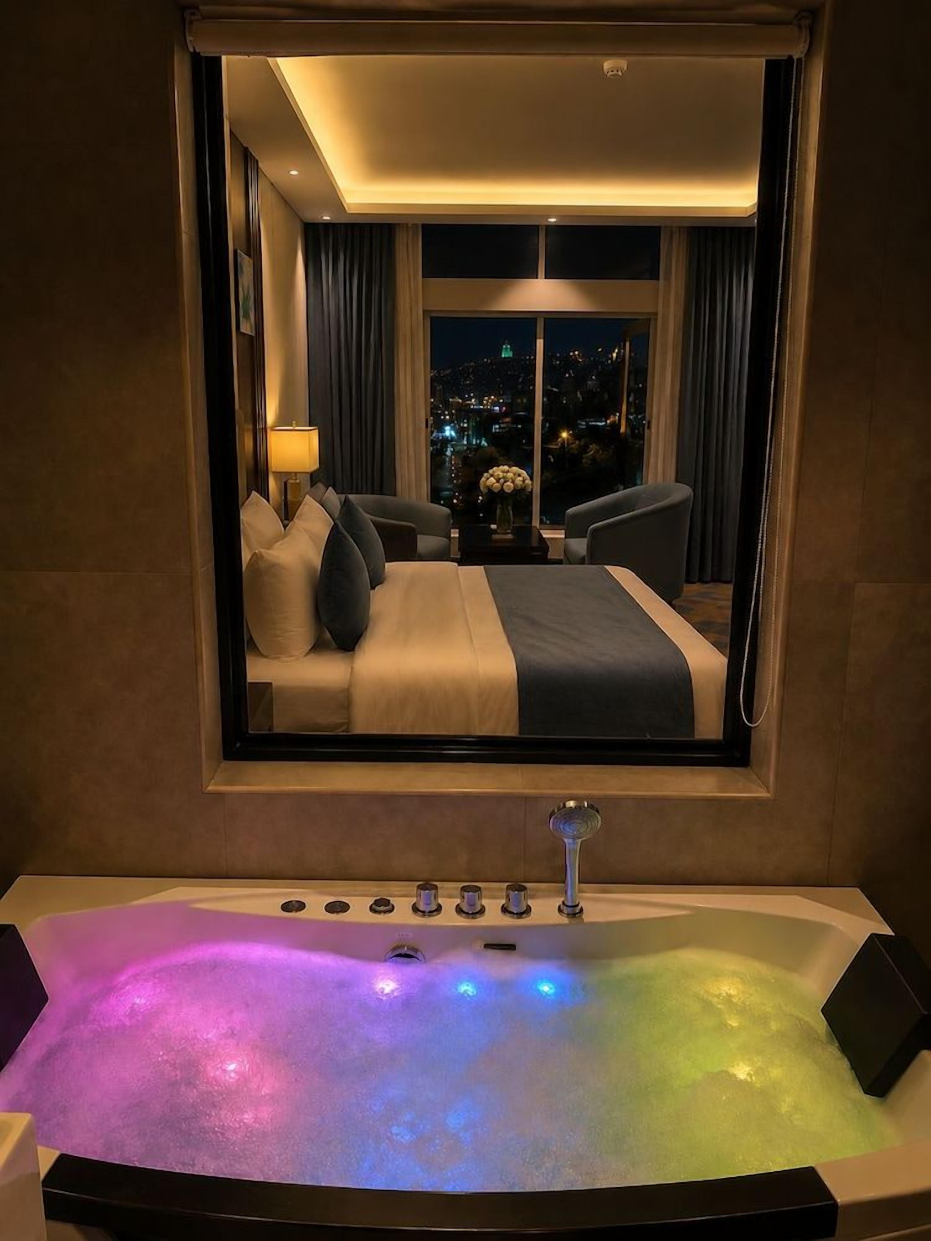 A hotel room with a large mirror reflecting a bed, a bathroom with a bathtub and control knobs, and a cityscape view through the window.