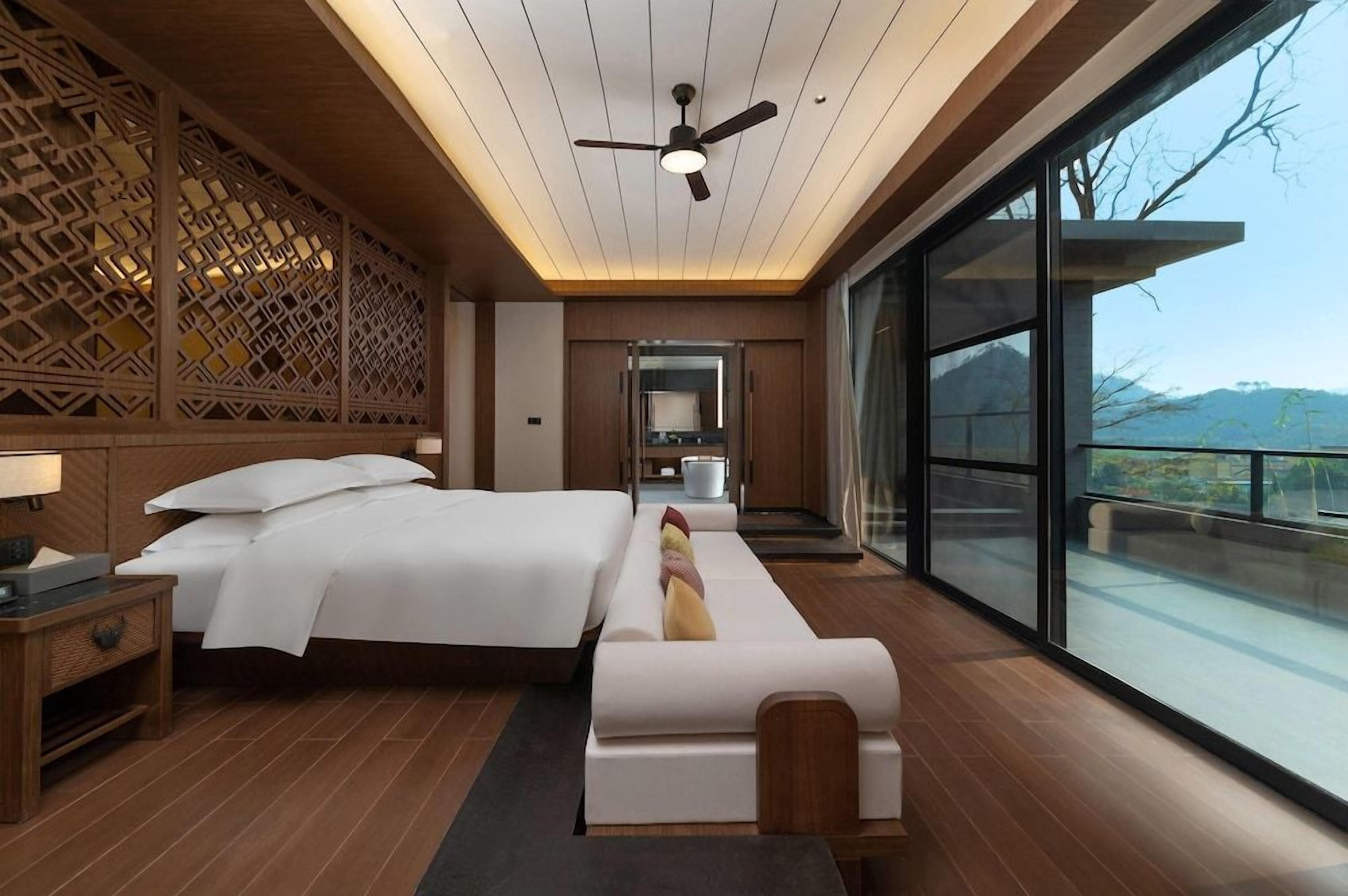 A modern hotel room with a large bed, a sofa, a ceiling fan, and a view of the outdoors.