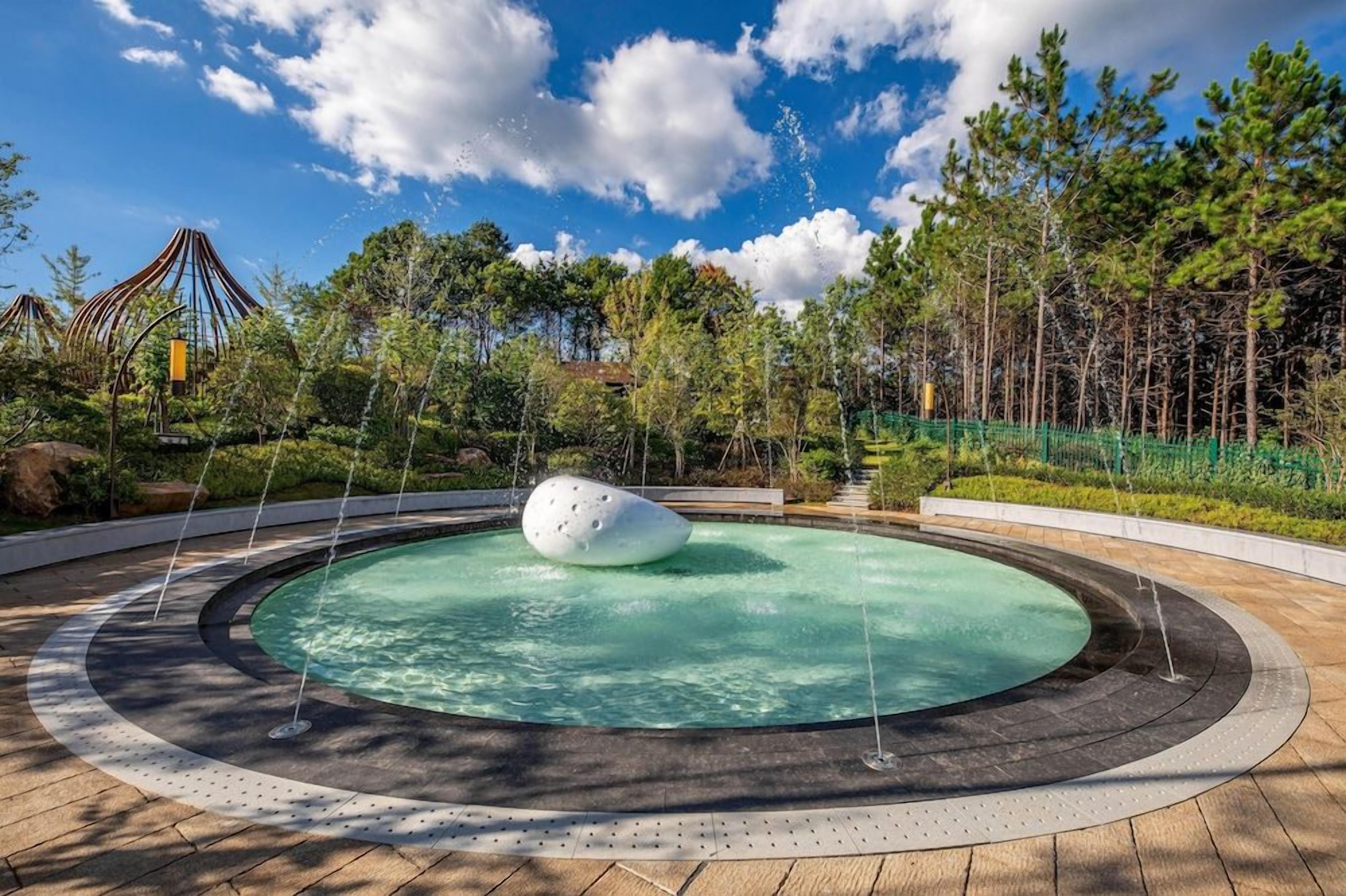 A circular water feature with a central sculpture, surrounded by a paved walkway and greenery.