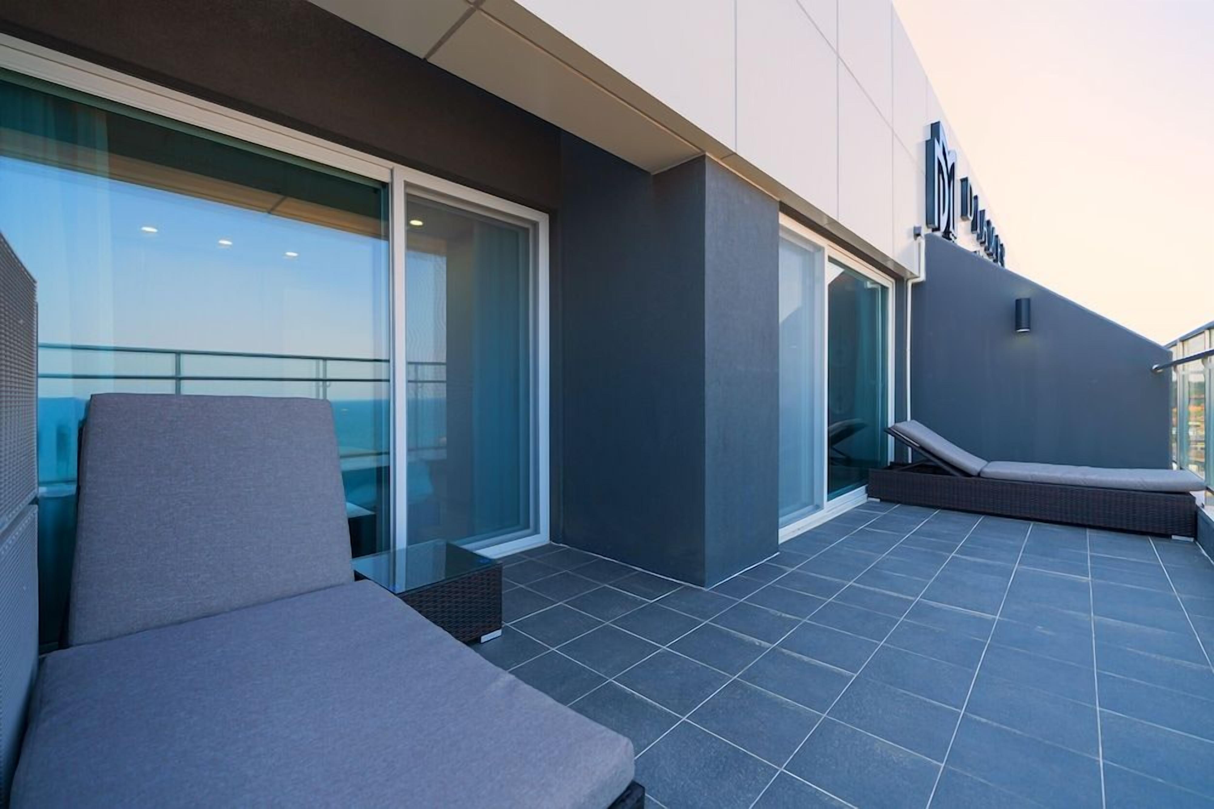 A modern outdoor lounge area with a glass balustrade, a cushioned seating area, and a clear view of the sky.