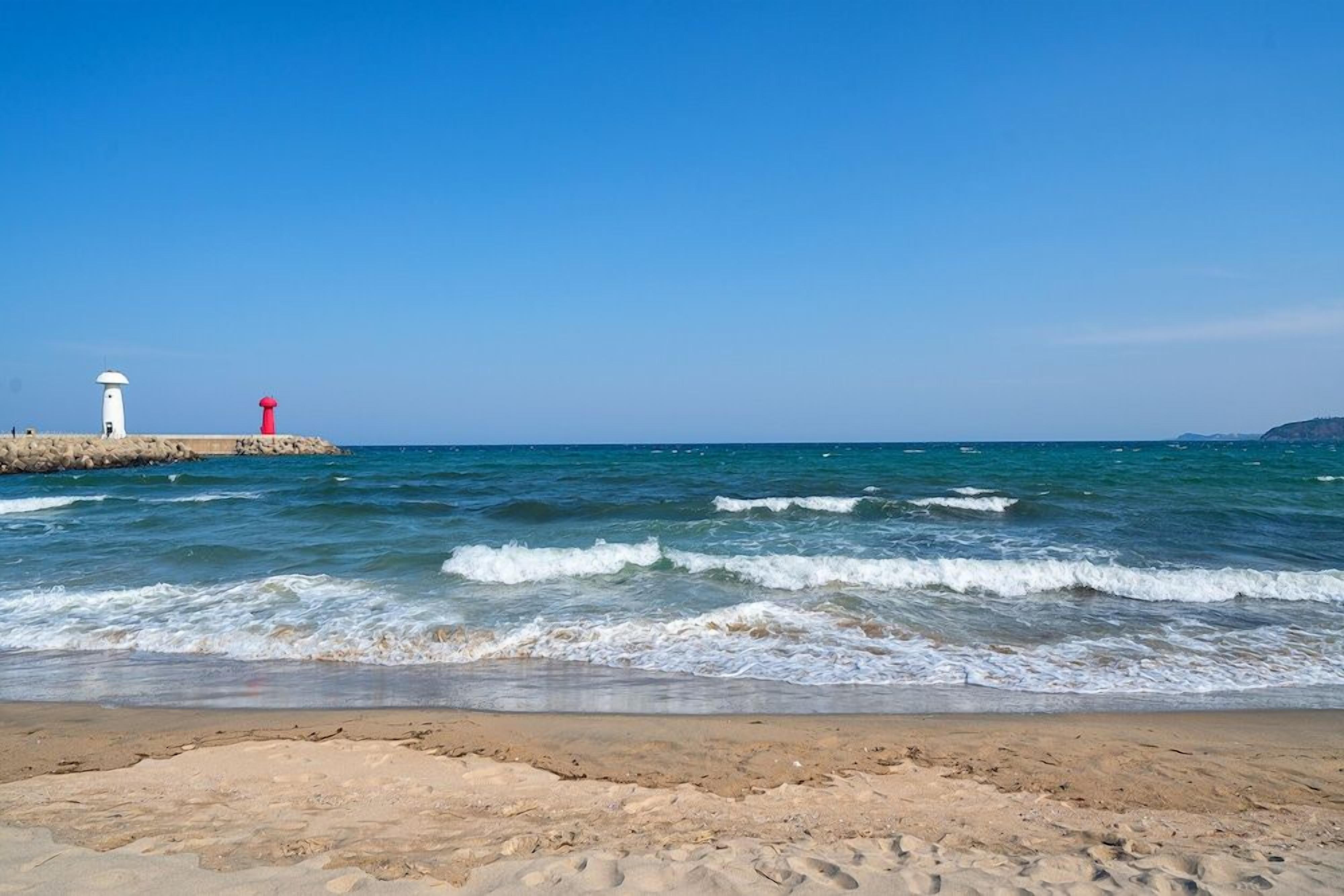 A beach with a white and red lighthouse, clear blue sky, and calm sea.