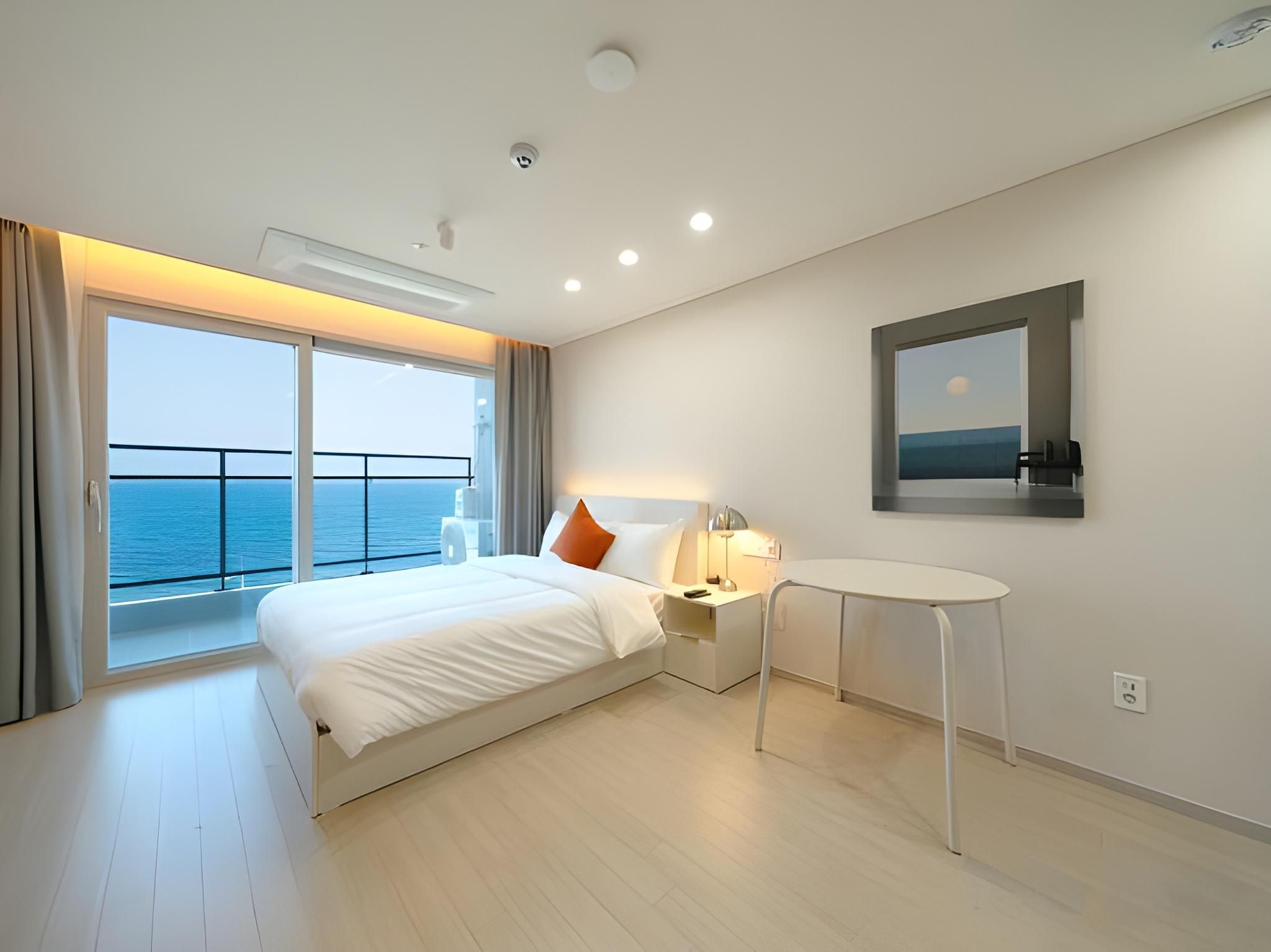 A modern hotel room with a large bed, a bedside table, a small round table, a nightstand, a window with a view of the sea, and a mounted picture on the wall.