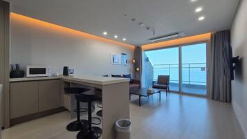 A modern kitchen with a bar area, a seating area with a sofa and chairs, and a large window offering a view of the sea.