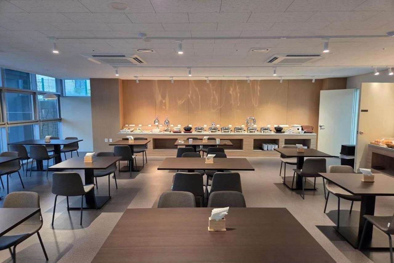 Una sala de conferencias moderna con mesas y sillas, una zona de cocina con electrodomésticos y un gran ventanal que proporciona luz natural.