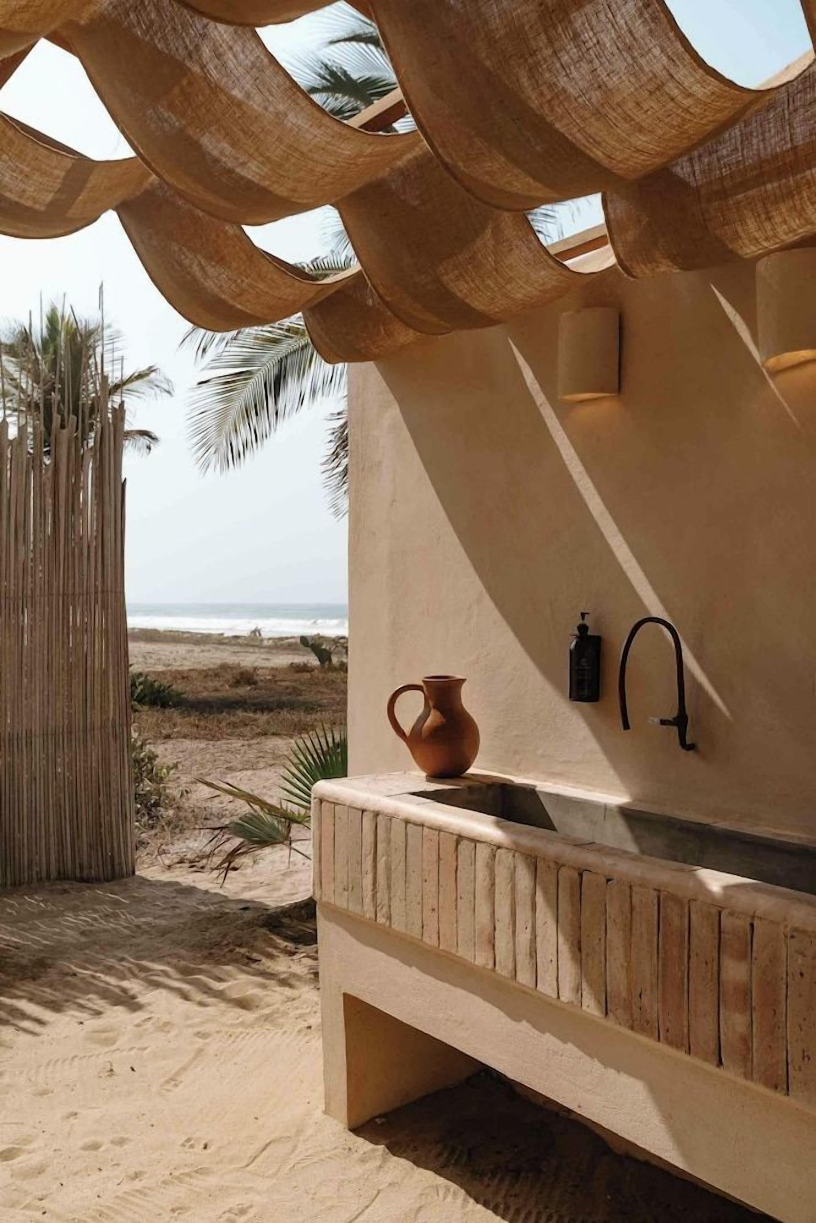 A beachside outdoor kitchen with a stone sink, a rustic wooden bench, and a woven shade structure.