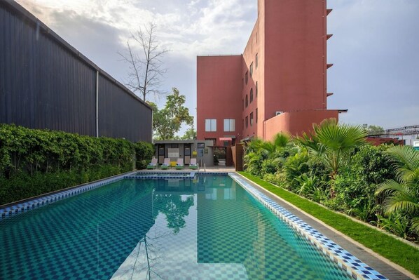 Une piscine à motifs à carreaux, entourée de verdure et bordée d’un bâtiment à la façade rouge.