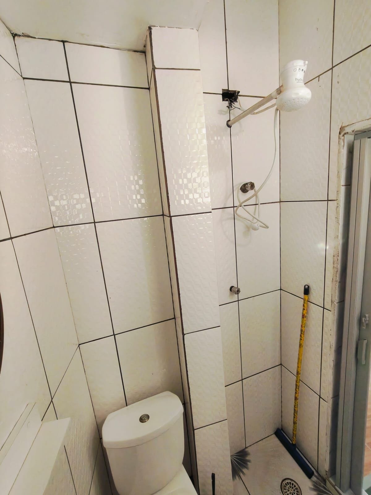 Casa de banho com paredes revestidas de azulejos brancos, sanita, chuveiro com duche manual e janela.