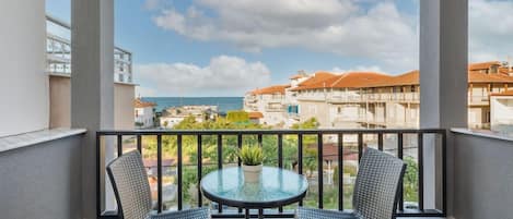 A balcony with two wicker chairs and a glass table, overlooking a coastal area with buildings and a clear sky.
