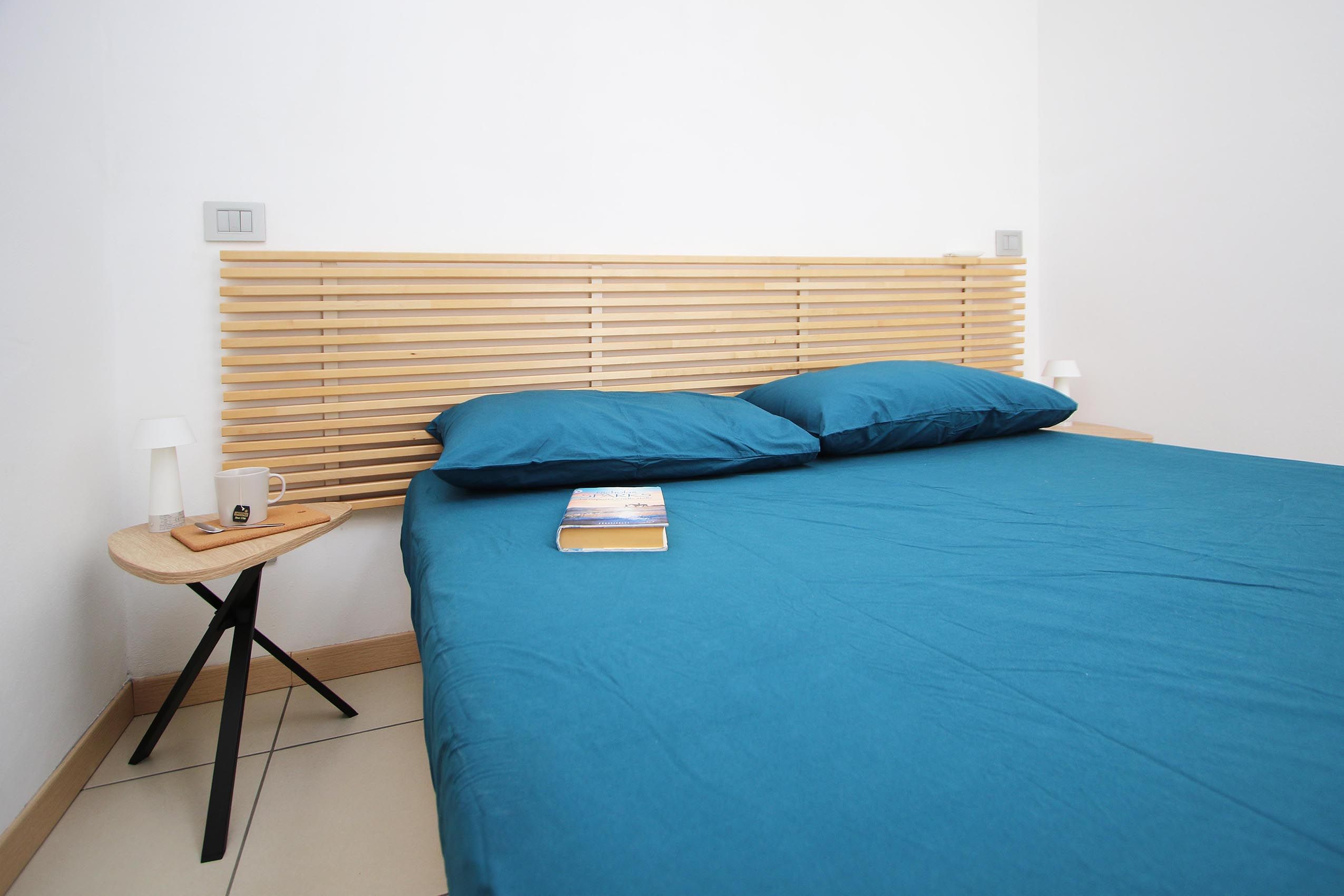 Ein Bett mit Holz-Kopfstück, einer blauen Matratze und zwei Kissen. Ein Nachttisch mit Lampe, einem Buch und einer Tasse.