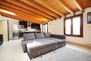 Ein modernes Wohnzimmer mit einem grauen Sofa, Holzbalkendecke, einer Küche im Hintergrund und einem großen Fenster.