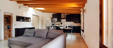 Una sala de estar moderna con un sofá gris, vigas de madera en el techo, una cocina al fondo y una estantería con libros y una planta.