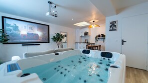 Ett modernt vardagsrum med en stor platt-TV, en sittgrupp med soffa, ett bubbelbad och ett köksområde i bakgrunden.