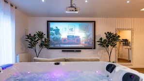 A modern living room with a large flat-screen TV mounted on the wall, a comfortable sofa, a hot tub with LED lights, and two potted plants.