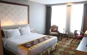 Quarto de hotel com uma cama, uma cadeira, uma mesinha e uma janela com cortinas.