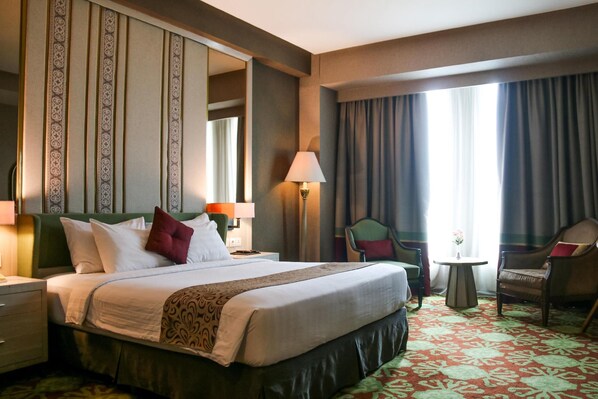 Quarto de hotel com uma cama grande, uma cadeira, uma mesinha e um abajur.