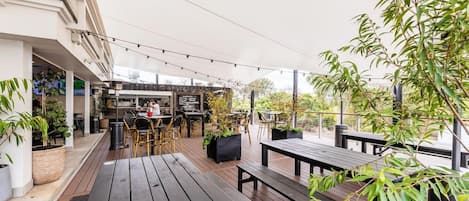 An outdoor patio area with wooden tables and chairs, a bar area, and potted plants.