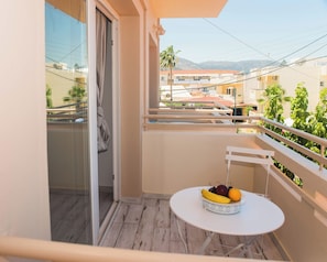 Un balcon avec une table sur laquelle se trouve un panier de fruits, une vue sur des immeubles et des palmiers, et un ciel dégagé.