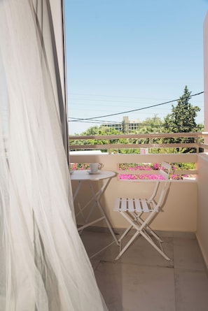 Uma varanda com mesa e cadeiras, uma janela com cortinas e vista para áreas verdes e edifícios.