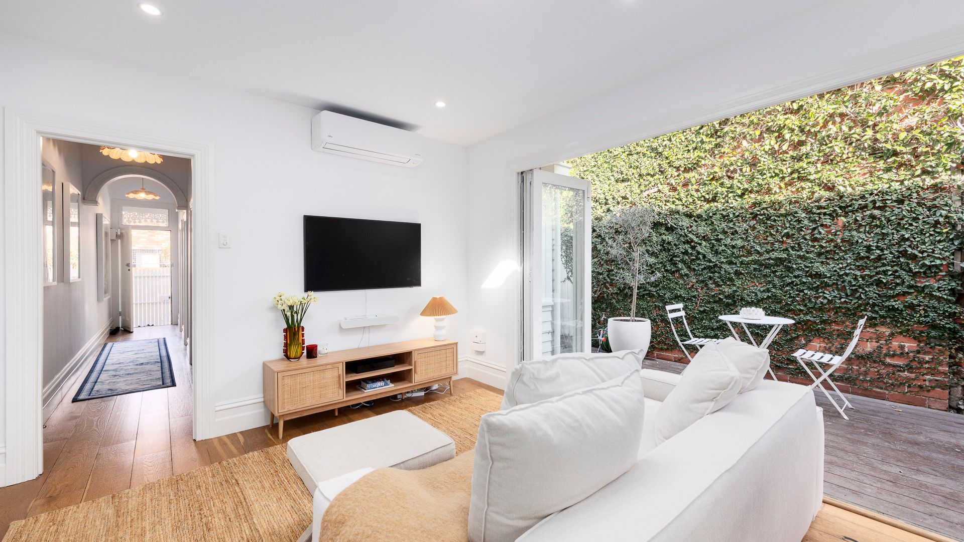 Una sala de estar moderna con un televisor de pantalla plana, un sofá, una mesa de centro y vistas a un seto verde exuberante a través de un ventanal.