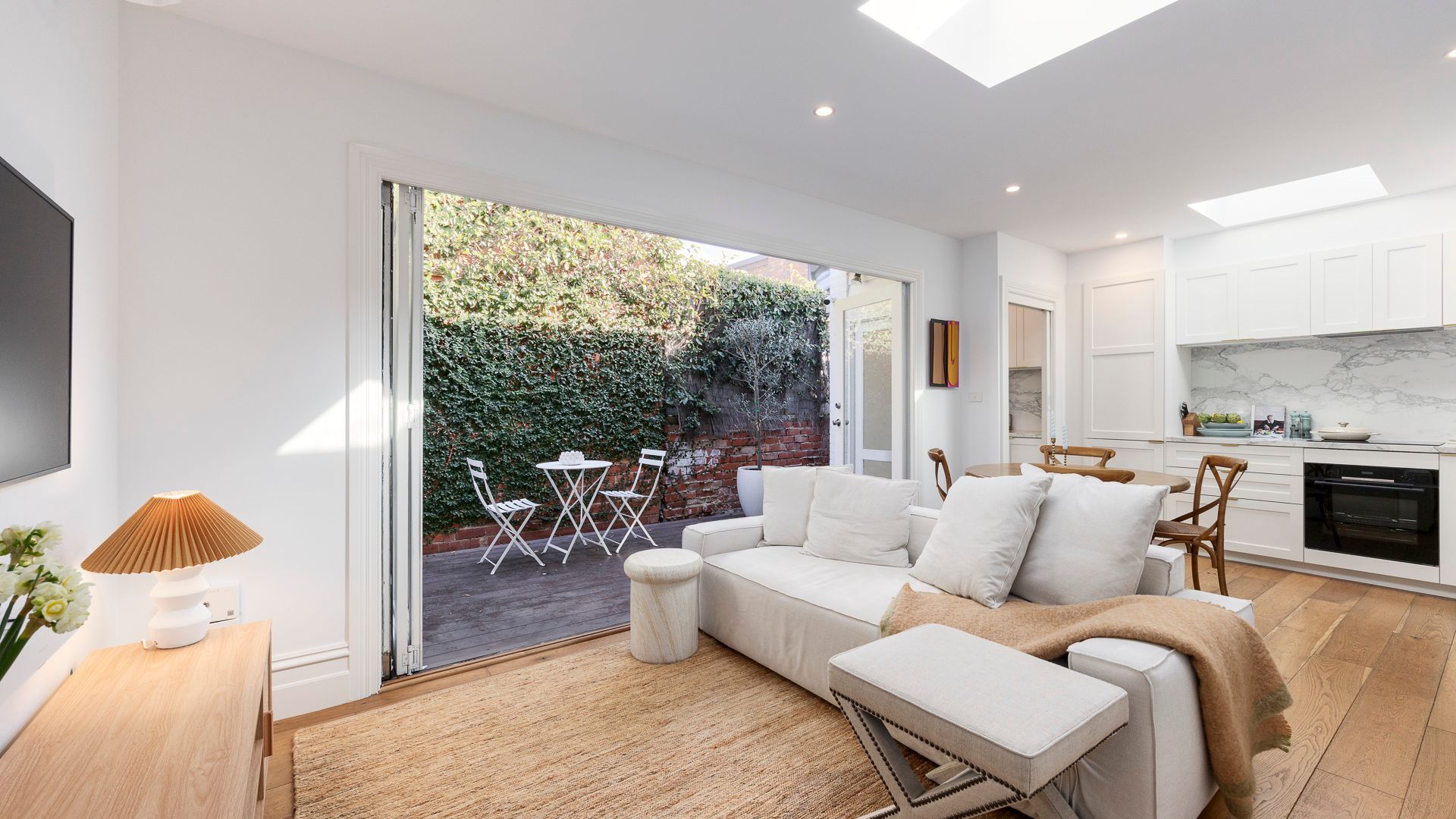 Ein moderner Wohnraum mit Sofa, Esstisch, St ühlen, einer Küchenzeile und Blick auf einen Garten durch eine Schiebetür.