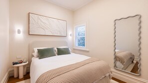 Ein Schlafzimmer mit Bett, Nachttisch, Wandbild, Fenster und einem großen Spiegel.