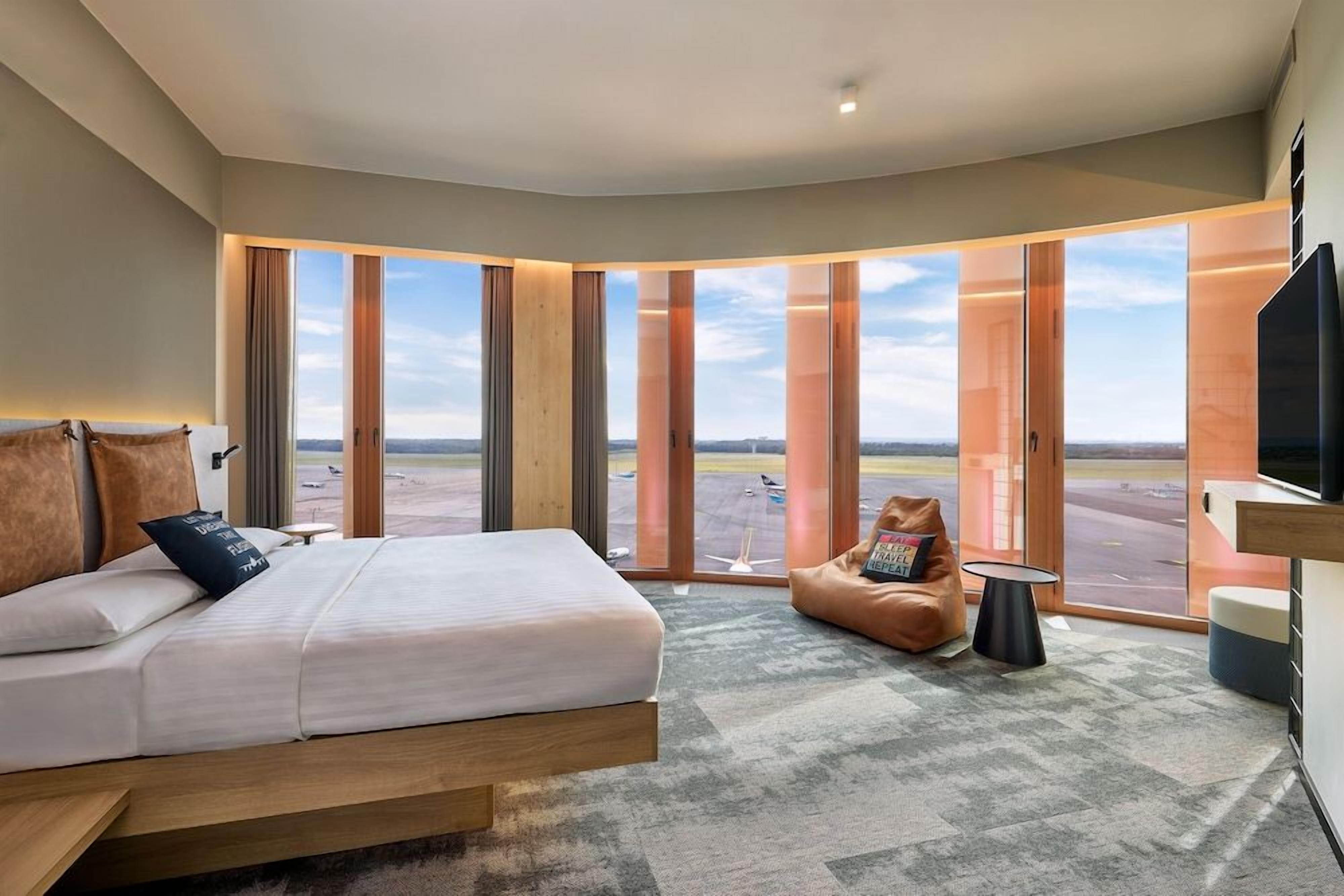 Quarto de hotel moderno com uma cama grande, um puff e uma televisão.