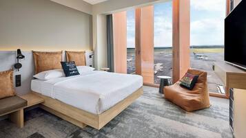 Ein modernes Hotelzimmer mit einem großen Bett, einer Sitzecke, einem Fernseher und Blick auf einen Flughafen.