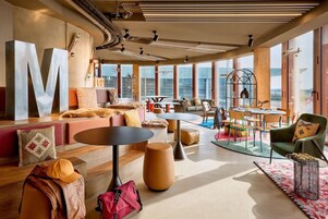 A modern lounge area with a large 'M' sculpture, multiple seating options, and a view of the cityscape through large windows.