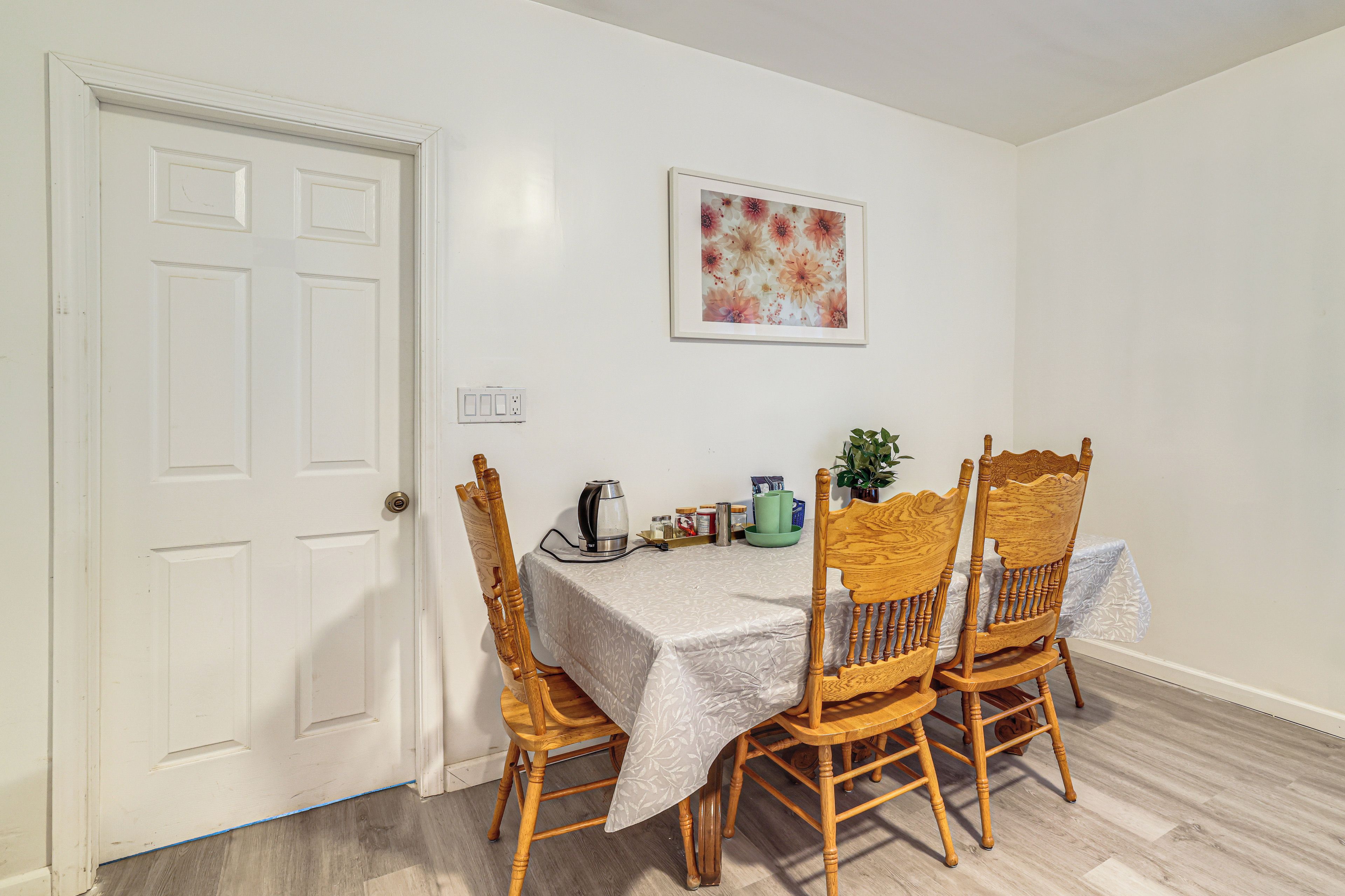 A dining area with a table set for four, surrounded by wooden chairs, a kettle, and a framed floral artwork on the wall.