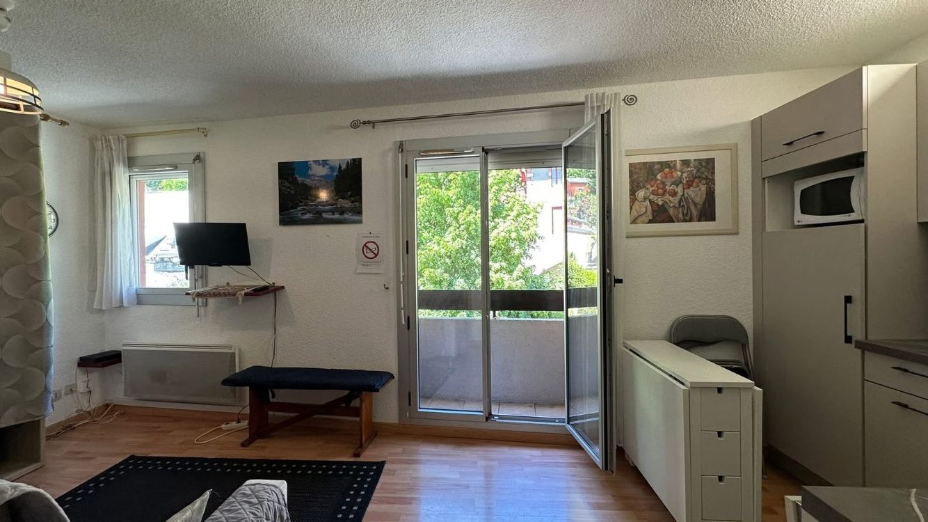 A room with a sliding glass door to a balcony, a bench, a television, a microwave, and a small cabinet.