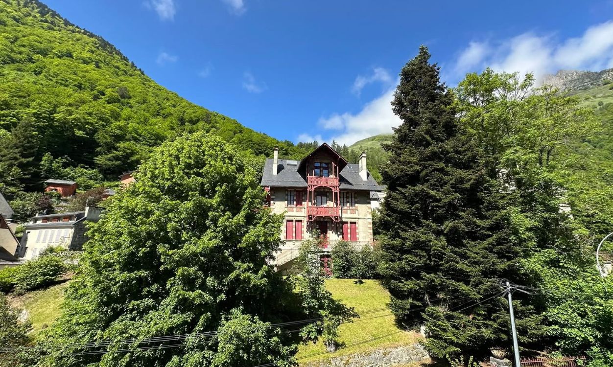 A traditional chalet with a balcony, surrounded by lush greenery and a mountain backdrop.