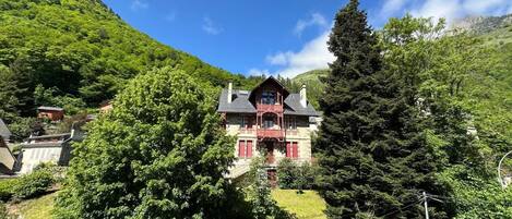 A traditional chalet with a balcony, surrounded by lush greenery and a mountain backdrop.