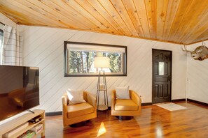 A modern living room with a wooden ceiling, a large window, two armchairs, a floor lamp, and a television. - Hotevilla Hideaway, Cozy Cabin Retreat (Flagstaff)