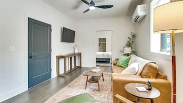 A modern living room with a sofa, a coffee table, a small table with a lamp, a ceiling fan, and a TV mounted on the wall.