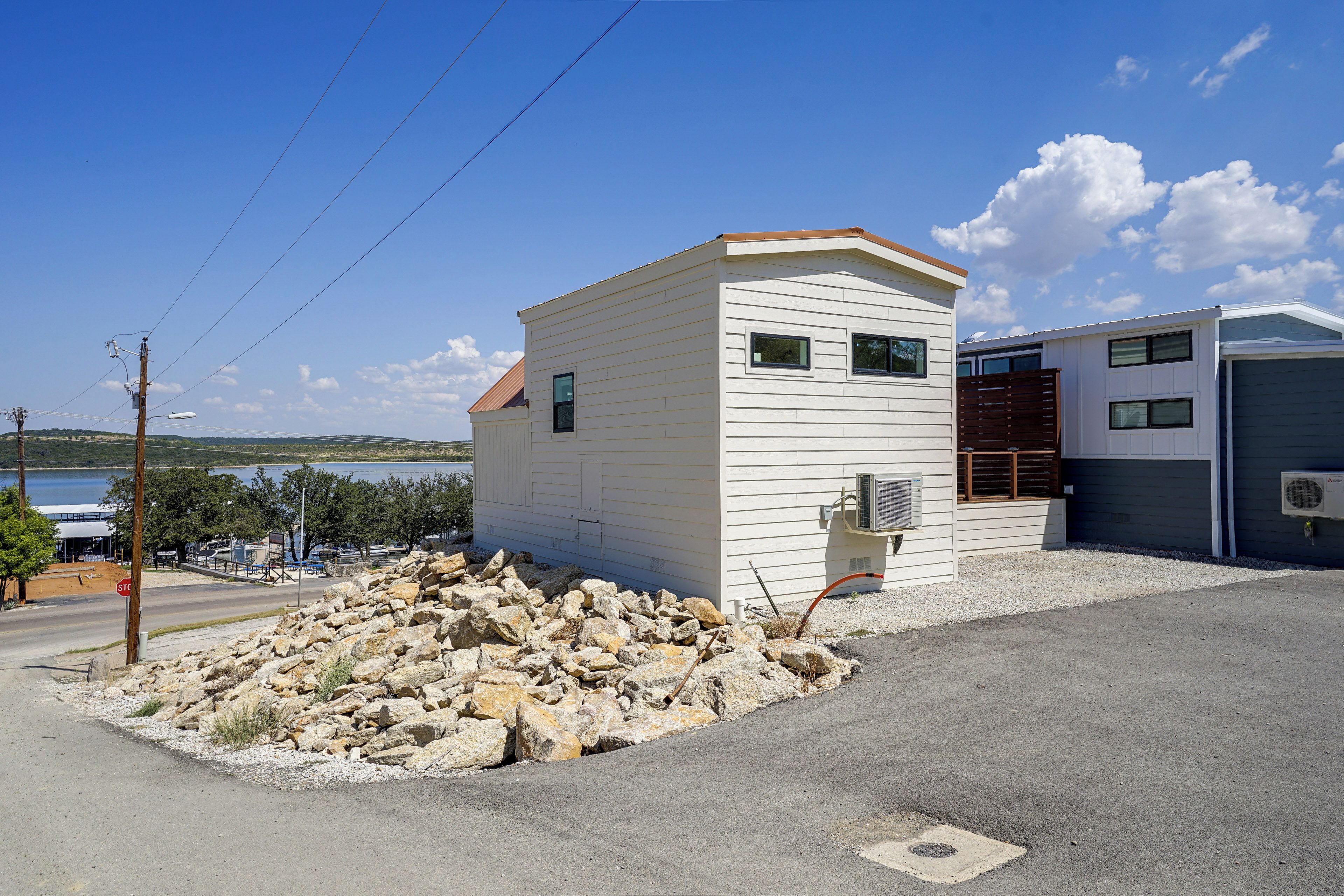 A small, single-story building with a white exterior and a brown roof, situated next to a body of water and a gravel driveway.