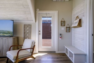 Una habitación con piso de madera, un banco blanco, un televisor sobre un mueble bajo y una puerta que da a vistas del exterior.