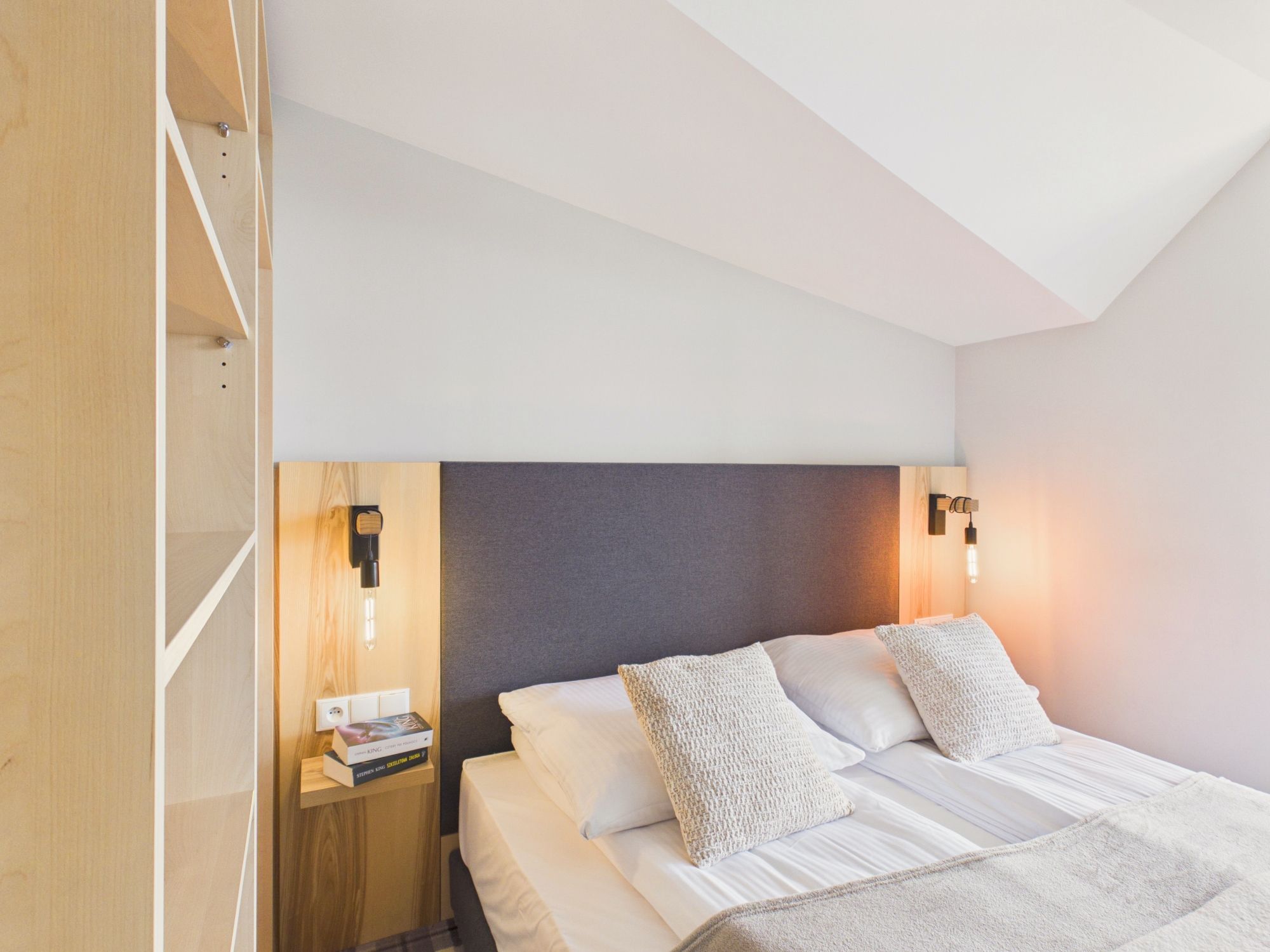 Un dormitorio con cama, almohadas, una mesita de noche, un libro e iluminación en la pared.