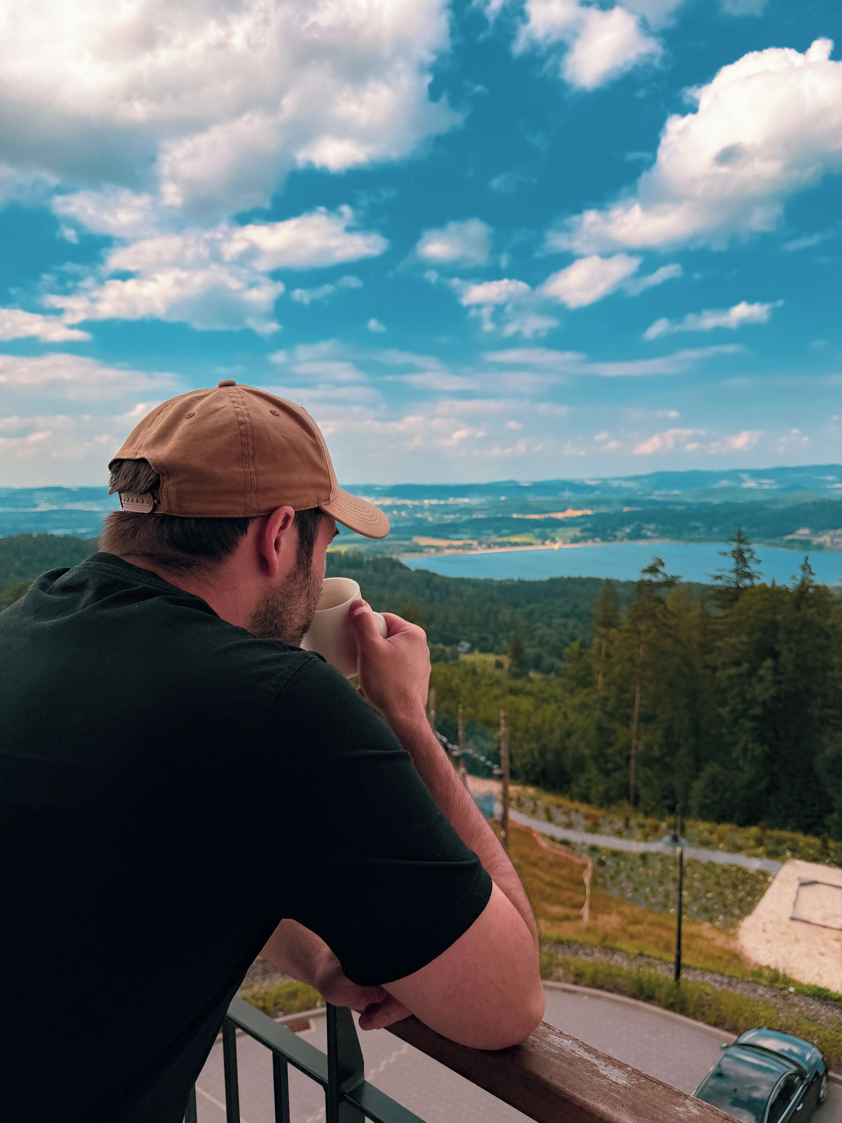 Una persona apoyada en la barandilla de un balcón, contemplando un paisaje pintoresco con un lago y colinas boscosas.