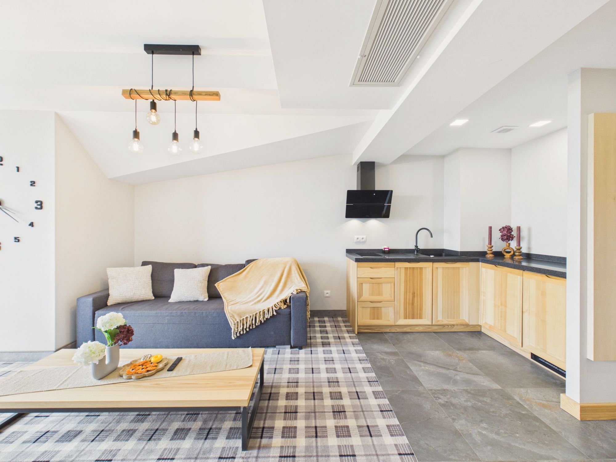 Un apartamento moderno con una sala de estar que incluye un sofá, una mesa de centro con aperitivos y una alfombra a cuadros. La cocina tiene armarios de madera y una encimera negra.