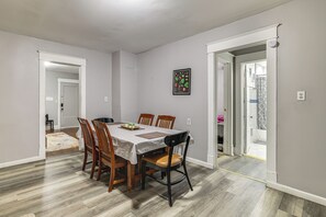 Uma sala de jantar com uma mesa posta para quatro pessoas, rodeada por cadeiras de madeira, uma impressão botânica emoldurada na parede e um banheiro visível por uma porta aberta.