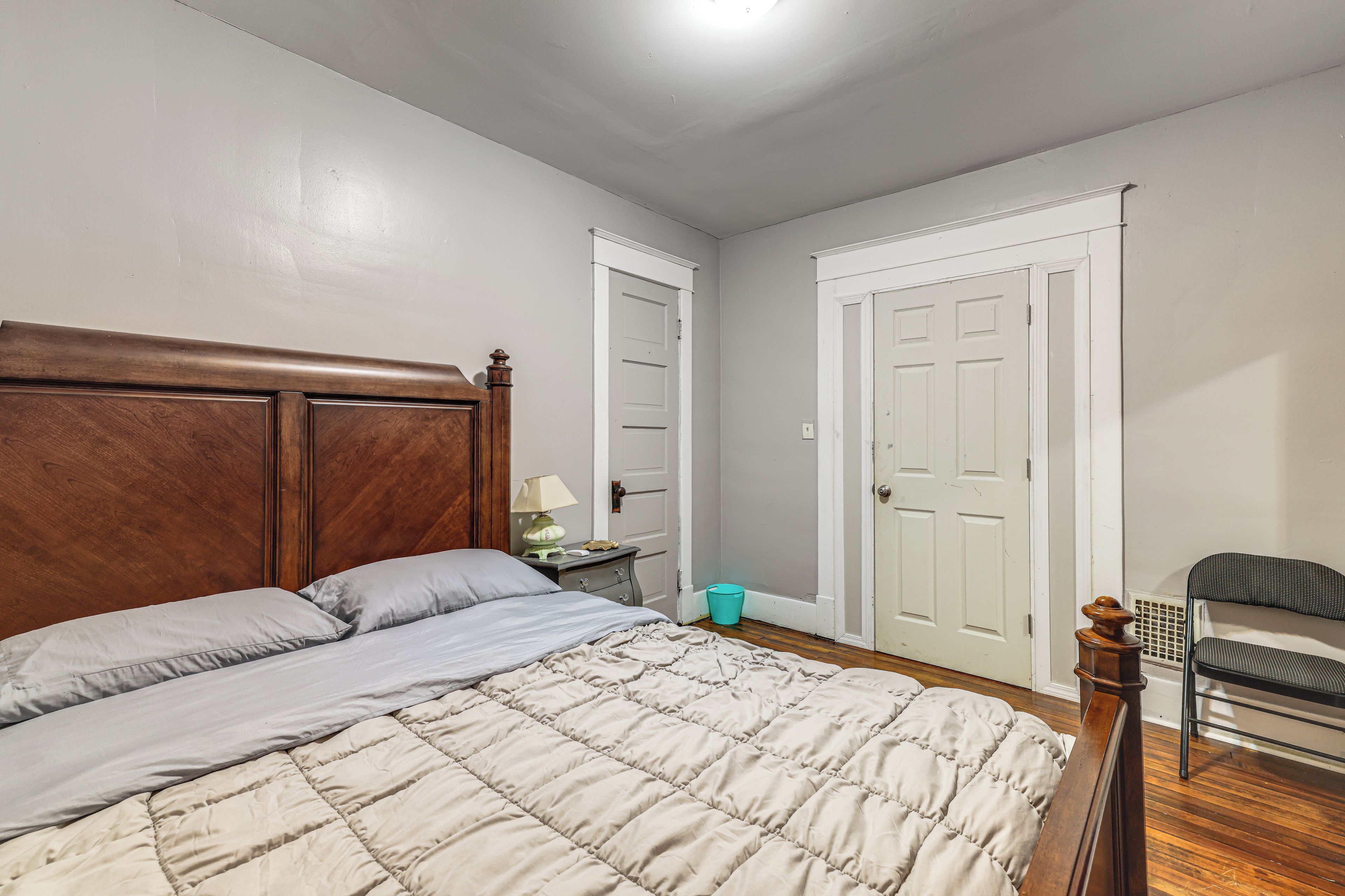 Quarto com cabeceira de madeira, cama com colcha bege, mesa de cabeceira com abajur e uma cadeira.