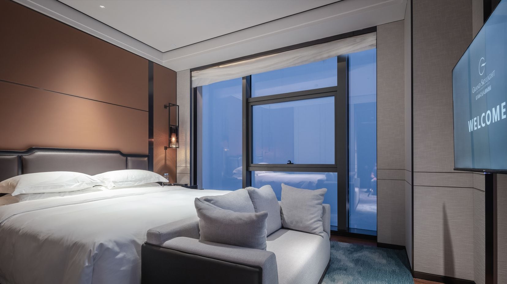A modern hotel room with a large bed, a sofa, a TV screen displaying 'WELCOME', and a view through the window.