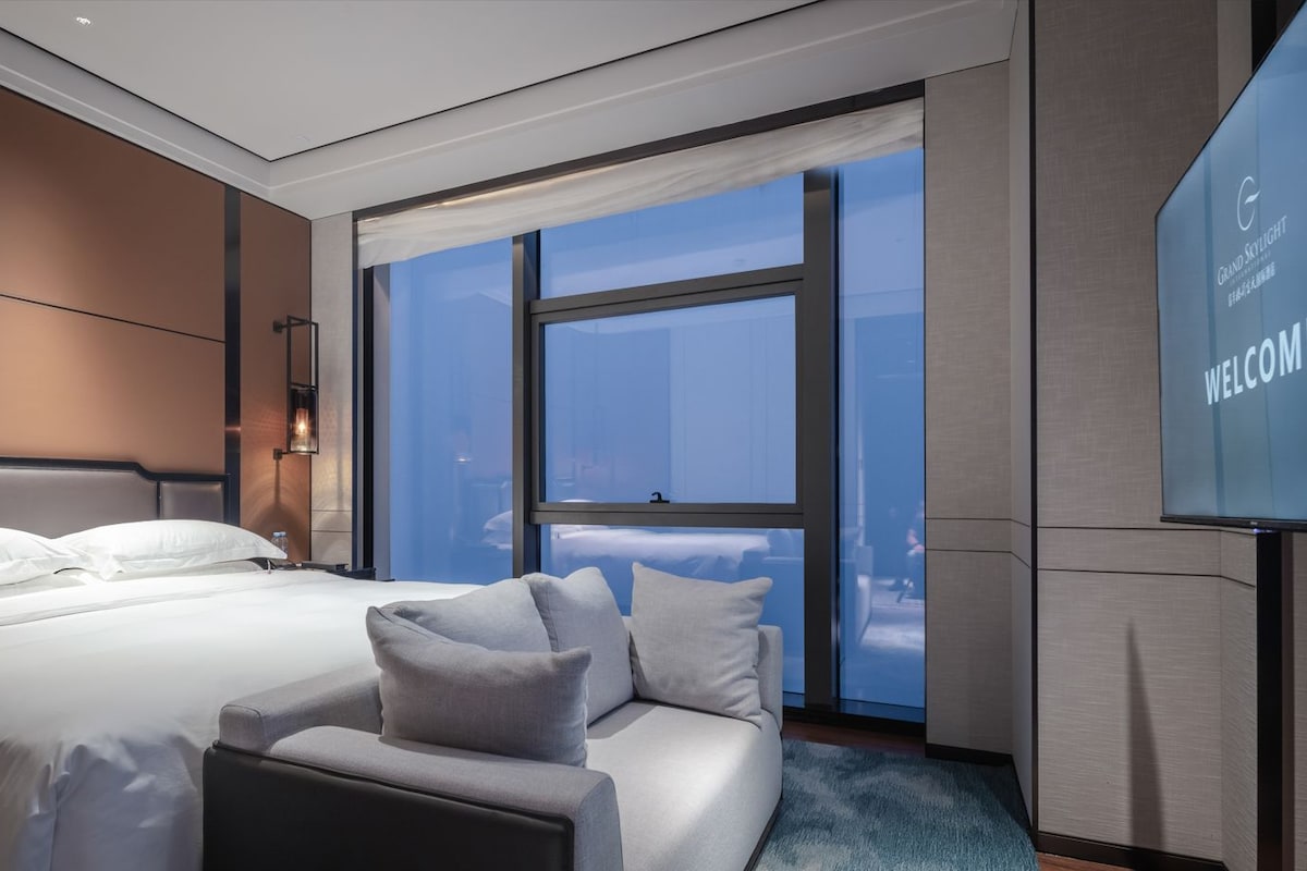 A modern hotel room with a large bed, a sofa, a TV screen displaying 'WELCOME', and a view through the window.