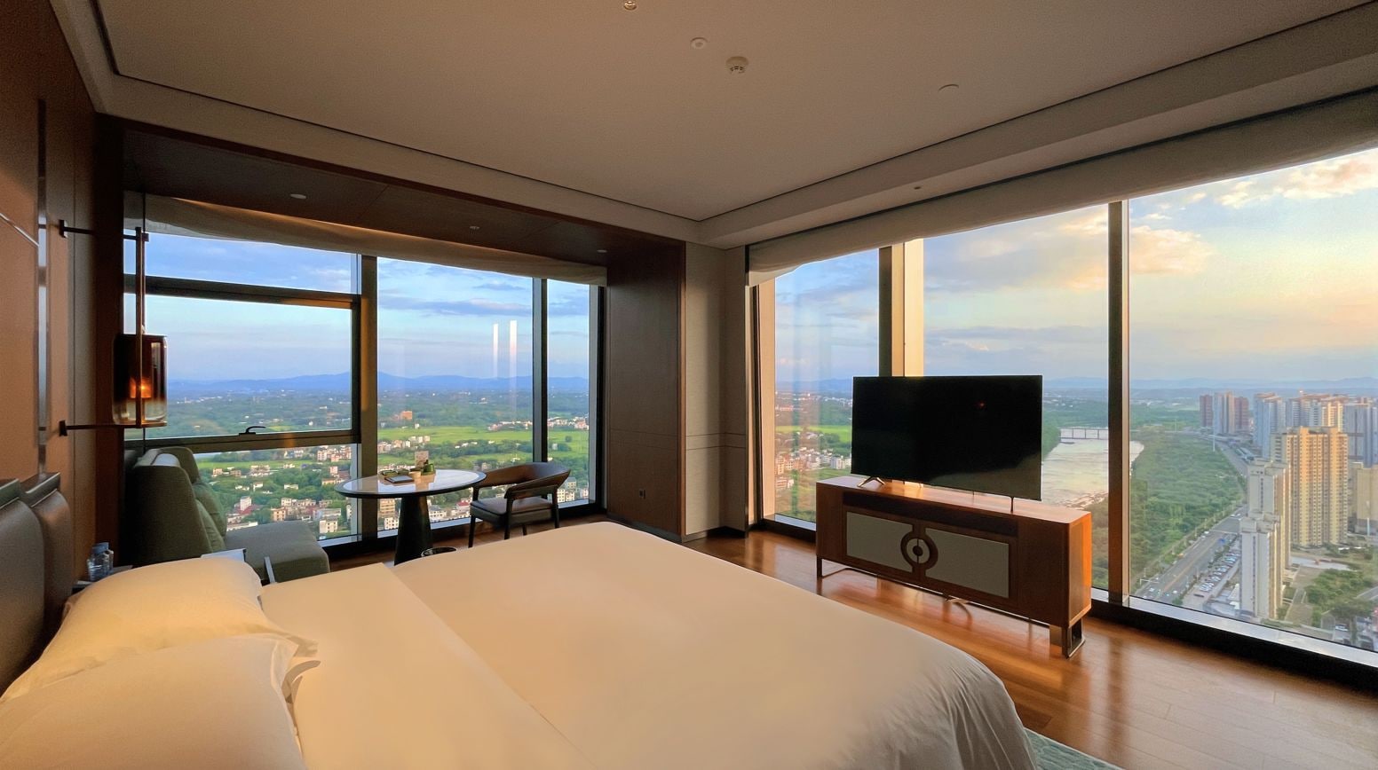 A hotel room with a large bed, a television, a small table, and a chair. The room has large windows offering a view of a cityscape.