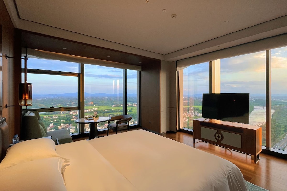 A hotel room with a large bed, a television, a small table, and a chair. The room has large windows offering a view of a cityscape.