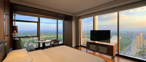 Habitación de hotel con una cama grande, televisión, una mesita y una silla. La habitación tiene ventanas amplias que ofrecen vistas de la ciudad.