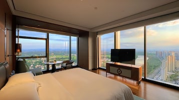 Habitación de hotel con una cama grande, televisión, una mesita y una silla. La habitación tiene ventanas amplias que ofrecen vistas de la ciudad.