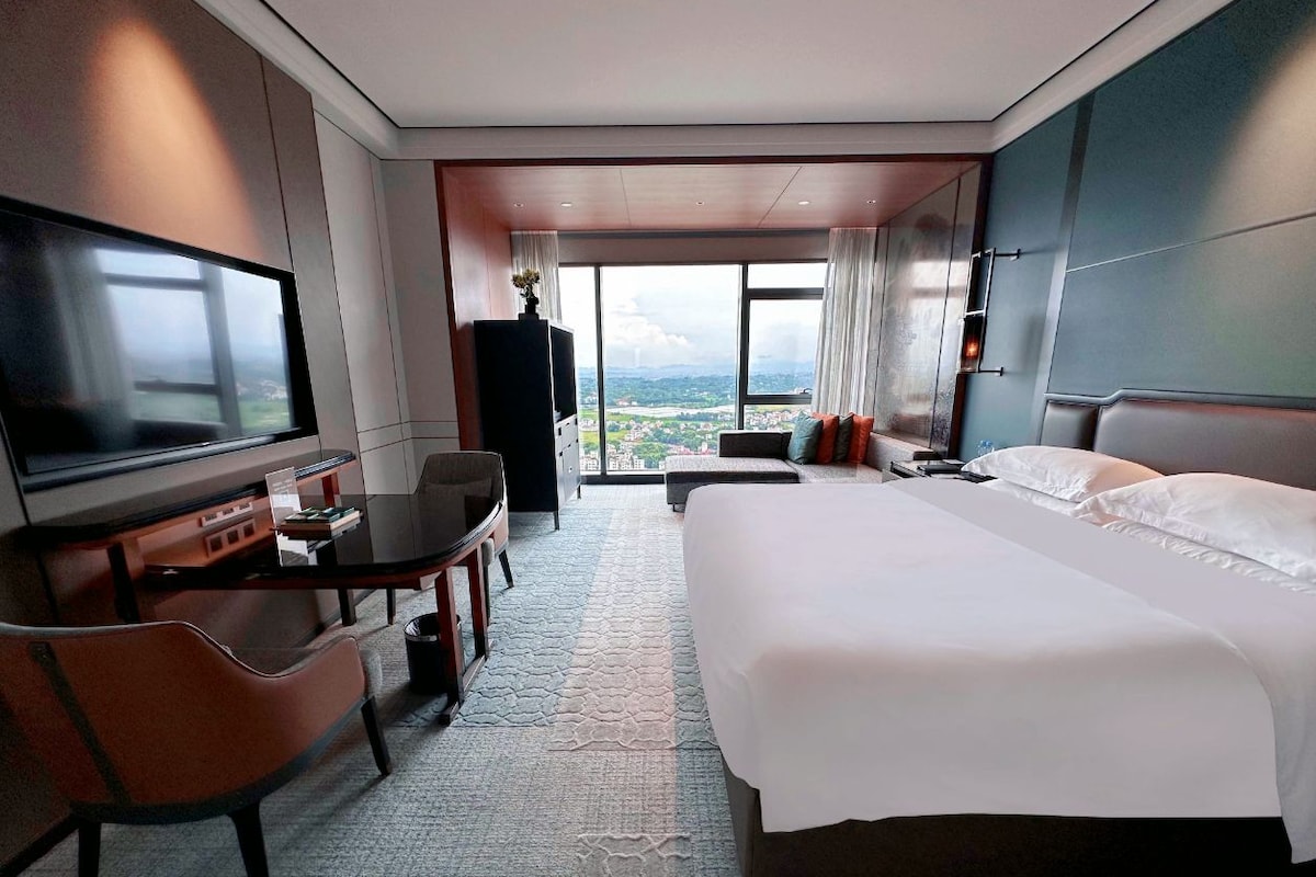 A hotel room with a large bed, a desk with a chair, a television, and a view of a cityscape through large windows.