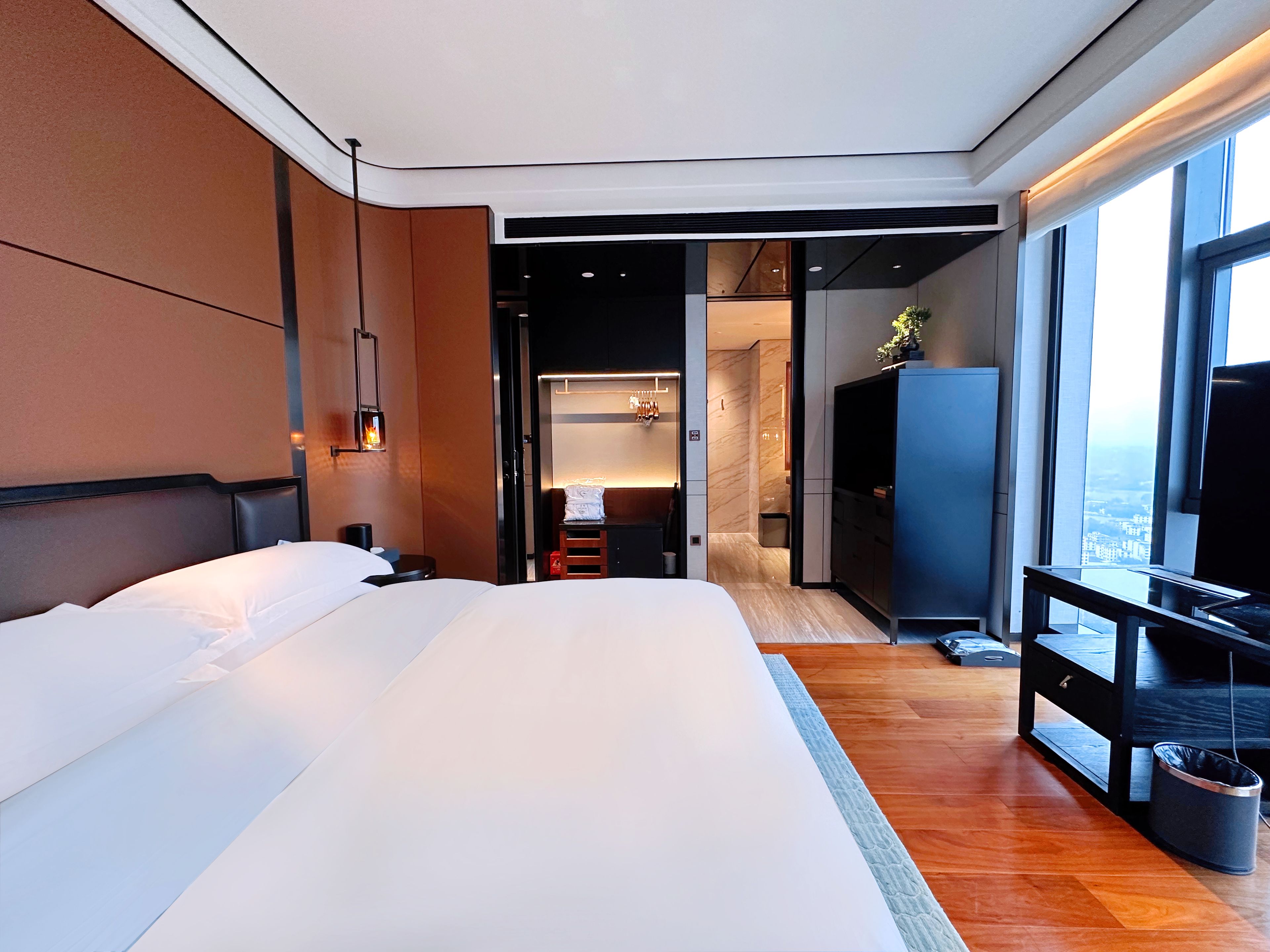 A modern hotel room with a large bed, a television, a desk, and a view of the city through the window.