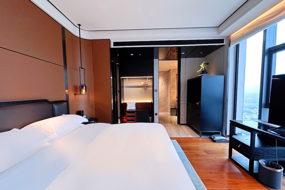 A modern hotel room with a large bed, a television, a desk, and a view of the city through the window.